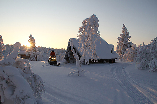 A snowmobile rider beside a snow-covered wilderness cabin at golden hour